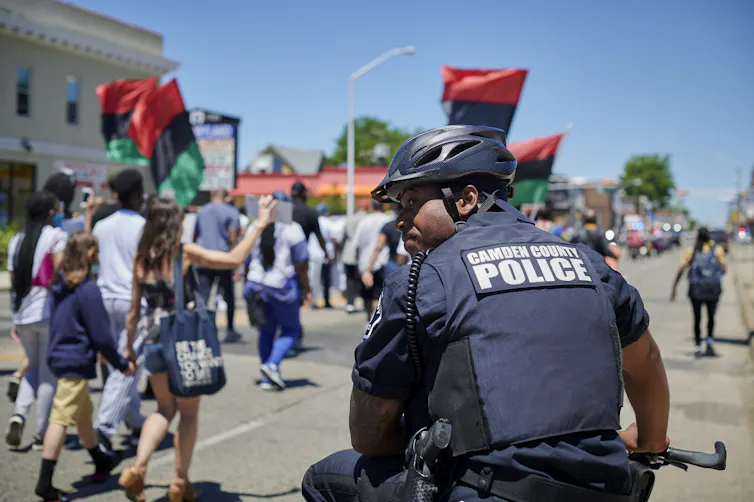 Police officer sits on bicycle as crowd of demonstrators waving red, black and green flags pass