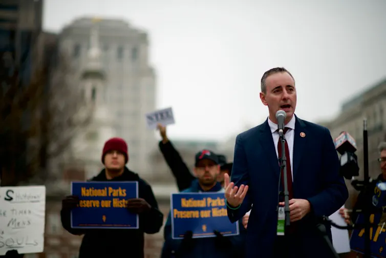 A man in a suit stands in front of a microphone outside.