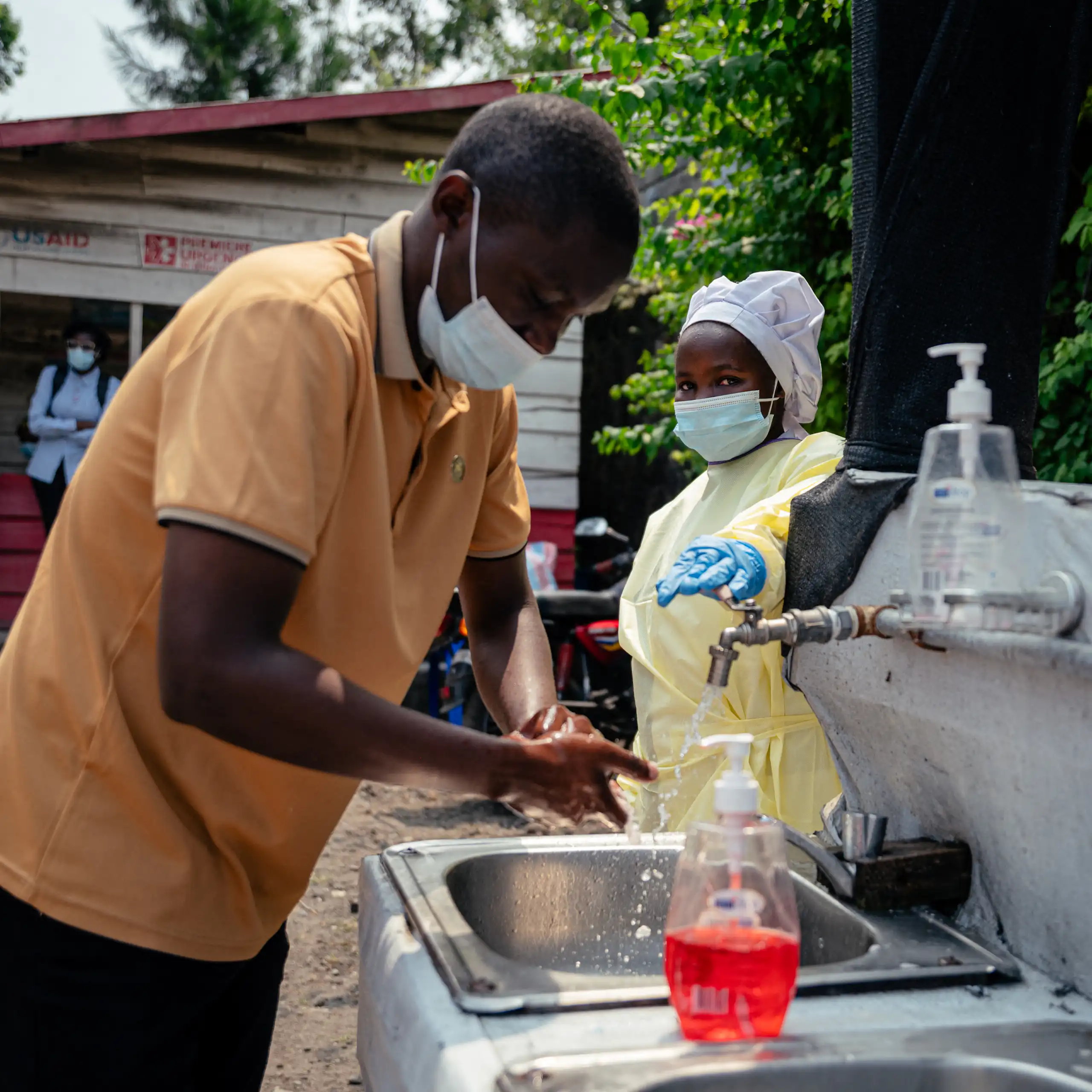 A person wearing a mask and protective clothing turns on an outdoor faucet for another masked person to wash their hands.