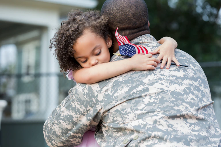 Child hugs soldier father holding small American flag during homecoming embrac