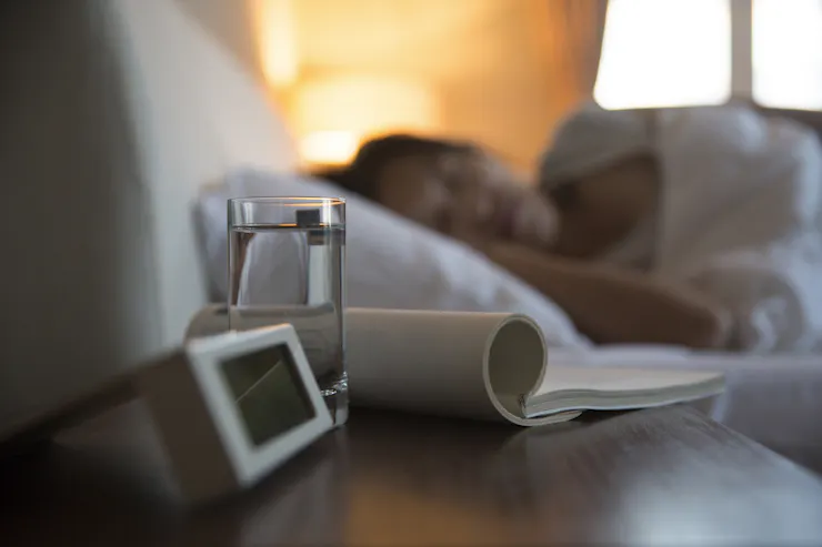 Close-up of a bedside table with an alarm clock, a glass of water and some reading material, with a person sleeping in the background.