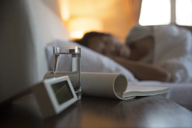 Close-up of a bedside table with an alarm clock, a glass of water and some reading material, with a person sleeping in the background.