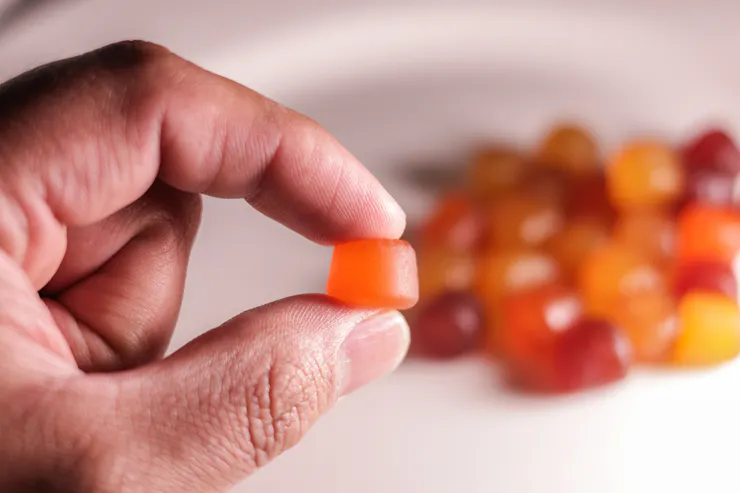 A hand holding up a single gummy in the foreground with a wide array of colorful gummies in the background.