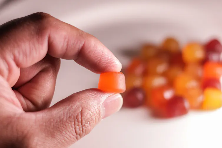 A hand holding up a single gummy in the foreground with a wide array of colorful gummies in the background.