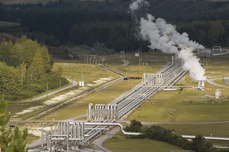 geothermal plant in New Zealand, steam rising from set of pipes with green grass and trees behind.