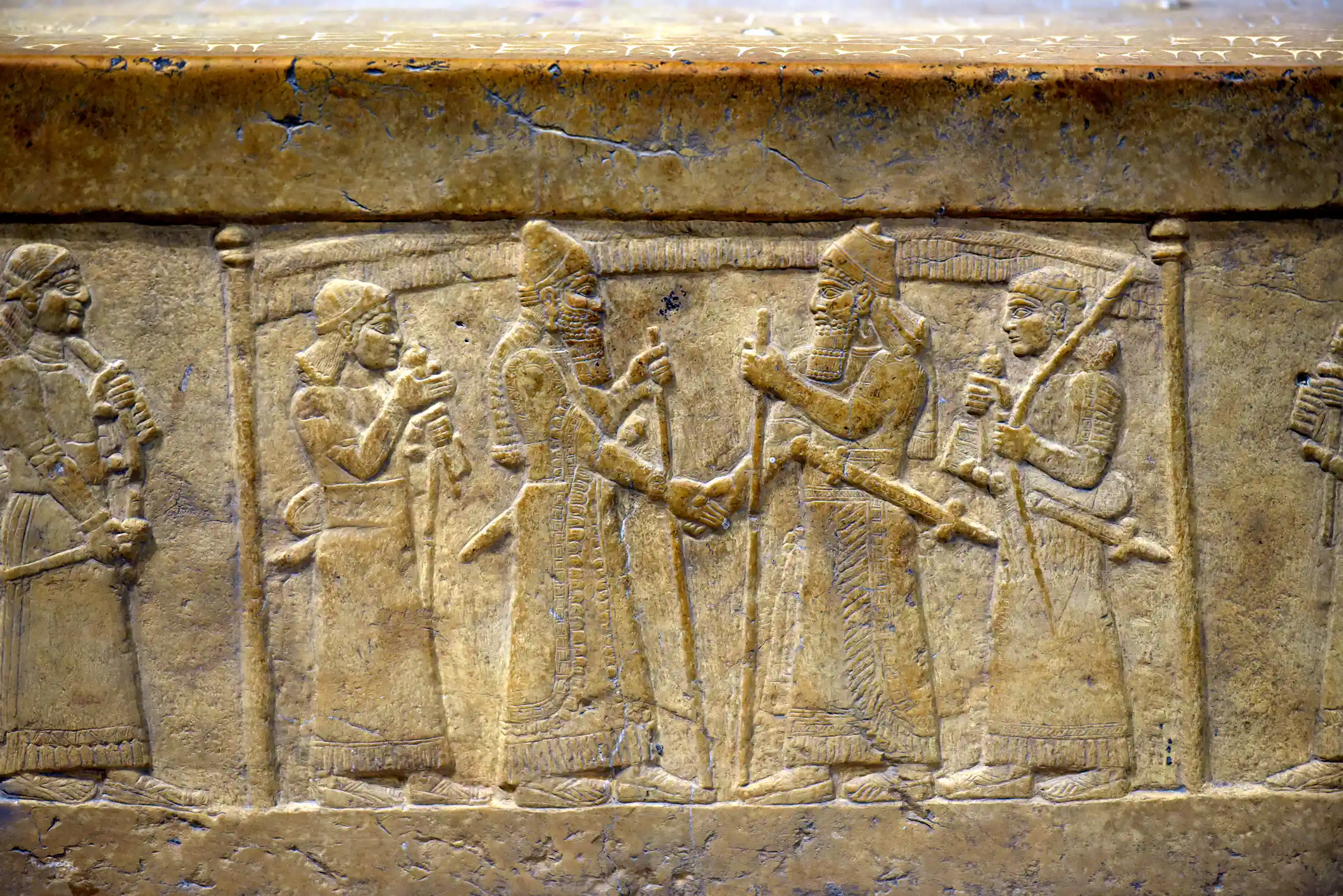A stone carving shows two men shaking hands. Both are surrounded by guards and stand below a fringed canopy supported by poles.
