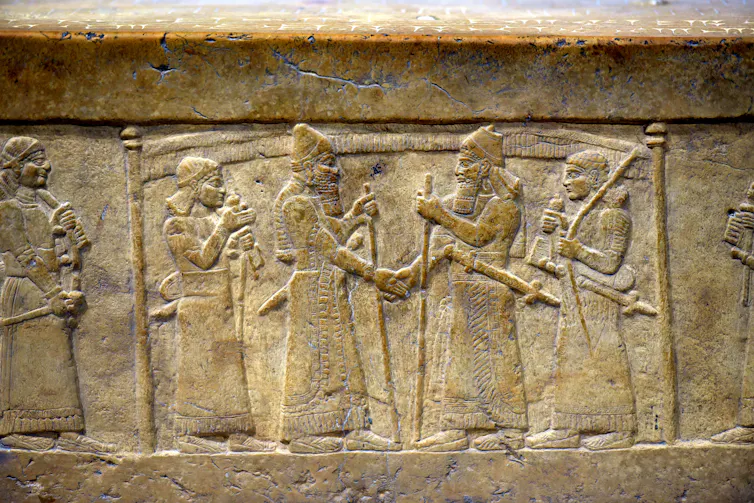 A stone carving shows two men shaking hands. Both are surrounded by guards and stand below a fringed canopy supported by poles.