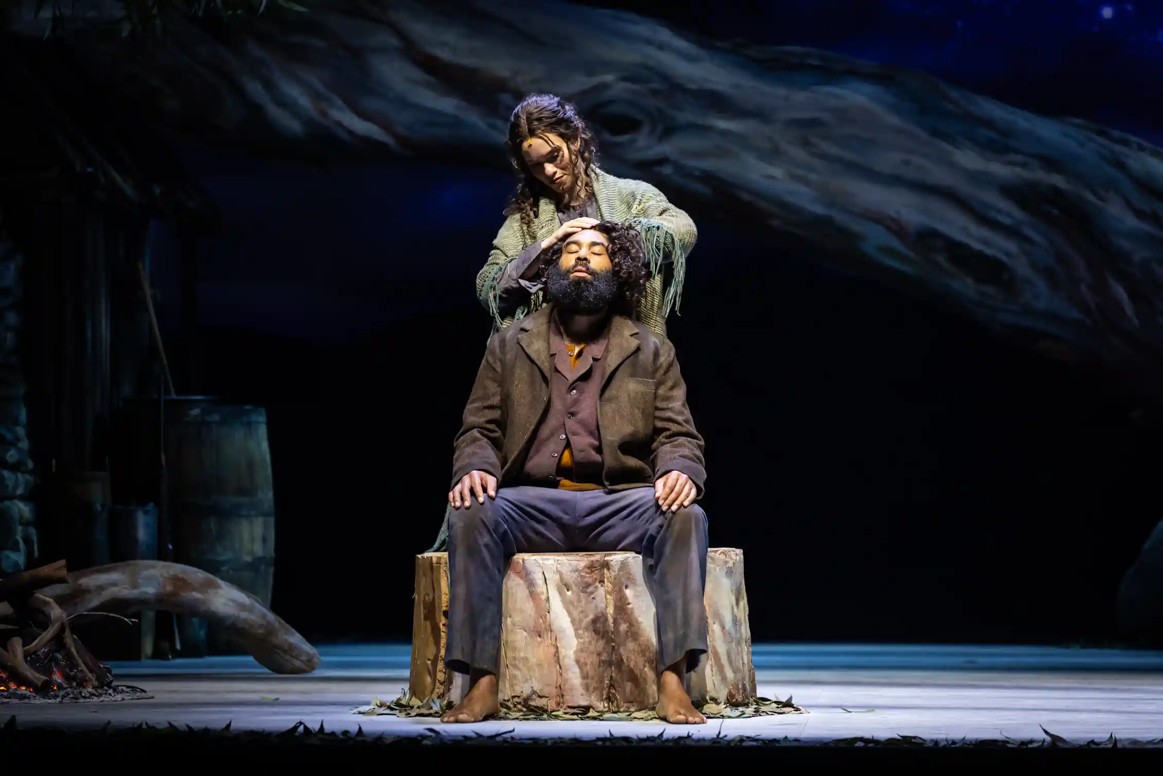 Production image: he sits on a tree stump; she stands behind.