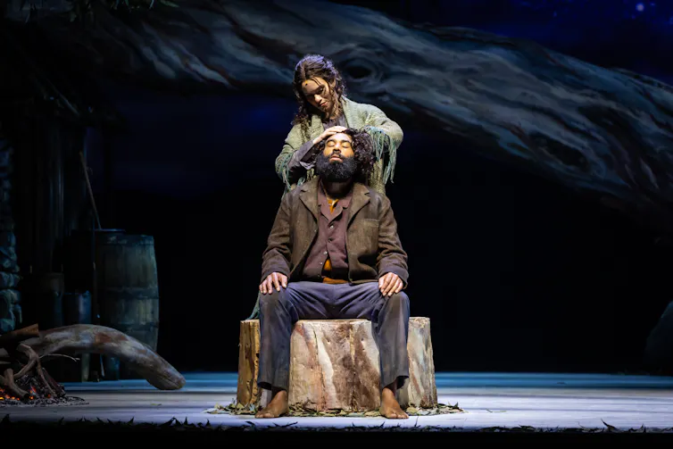 Production image: he sits on a tree stump; she stands behind.