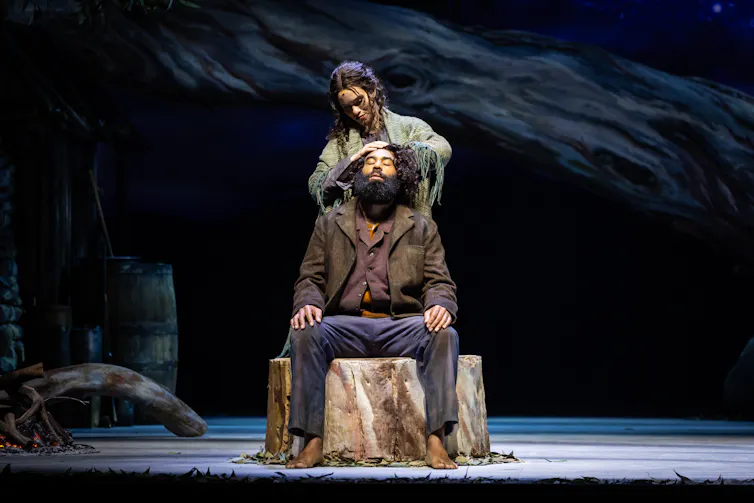 Production image: he sits on a tree stump; she stands behind.