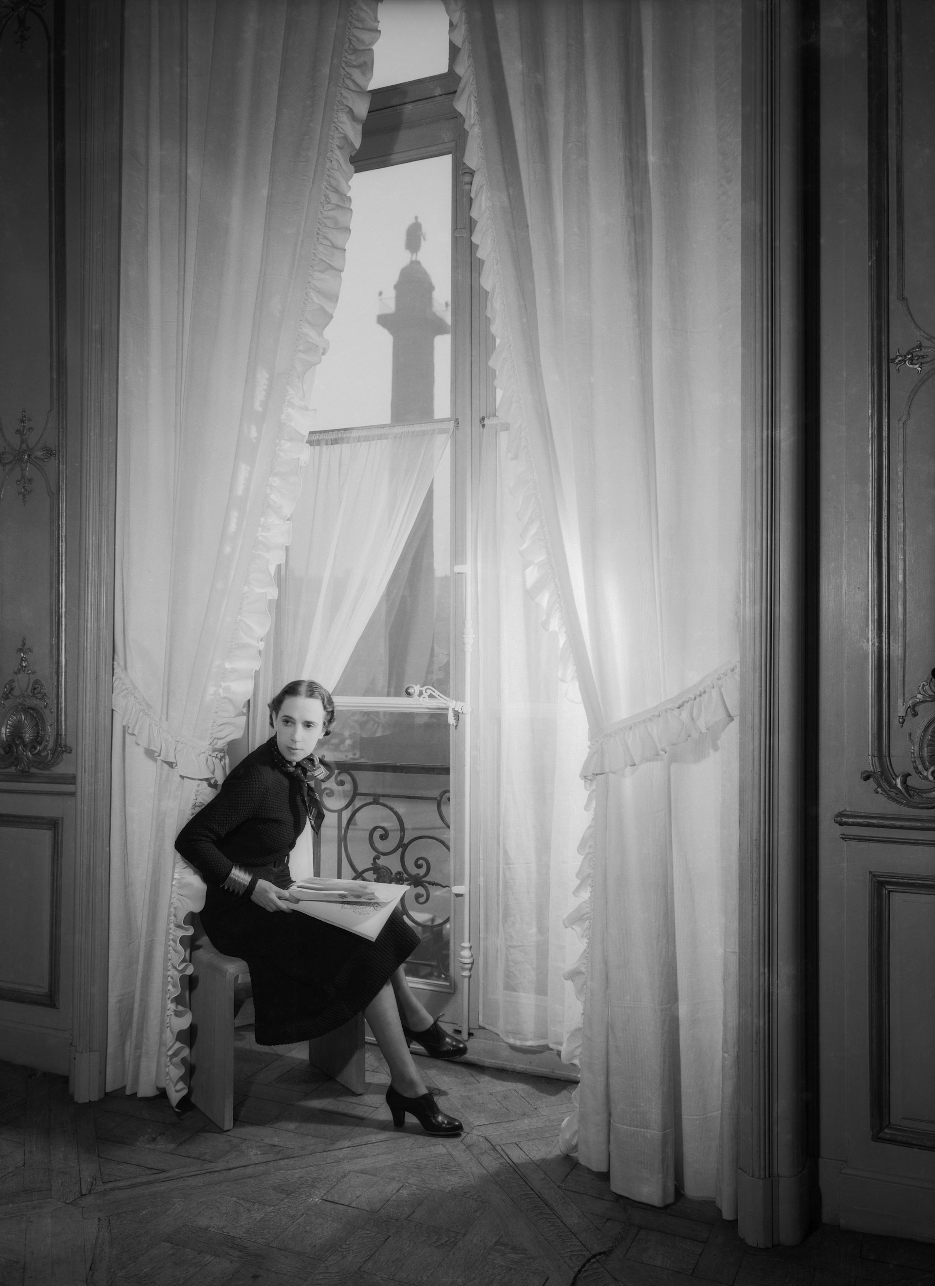 A black and white photograph of a stylish woman in a dark dress seated in front of a tall curtained window.
