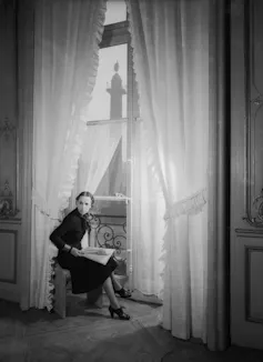 A black and white photograph of a stylish woman in a dark dress seated in front of a tall curtained window.