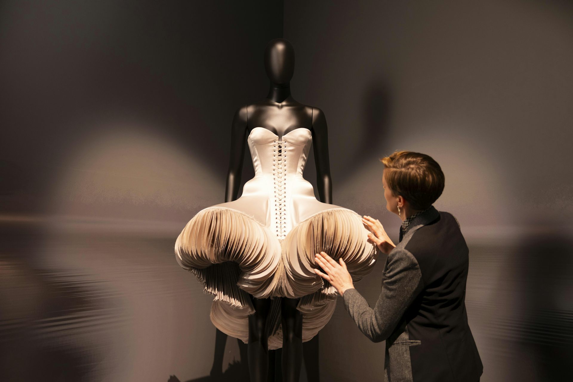 A conservator examines a blush coloured sculptural bodice dress in dramatic lighting.