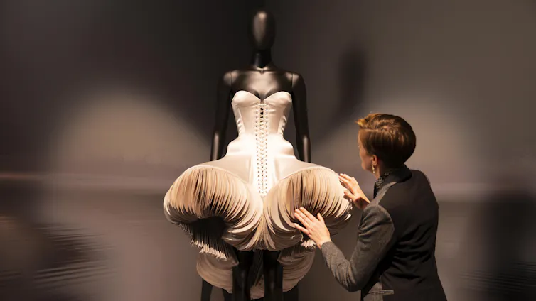 A conservator examines a blush coloured sculptural bodice dress in dramatic lighting.