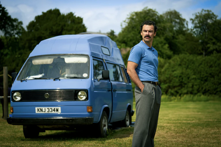 A man with a moustache wearing a blue shirt the same colour as the campervan next to him.