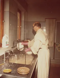 Photograph of British Benedictine priest Dom Sylvester Houédard standing in his cream robes washing up at a sink.