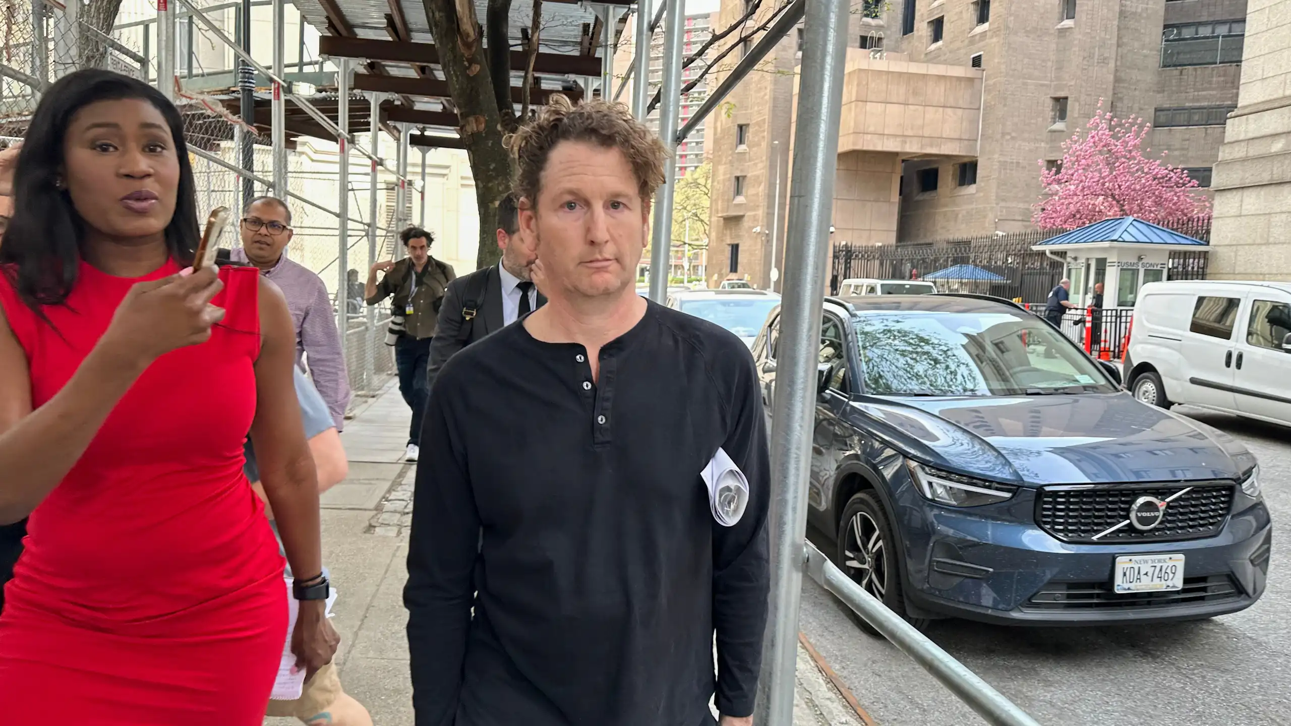 A man stares blankly ahead while walking and being videoed by a woman in a red dress.