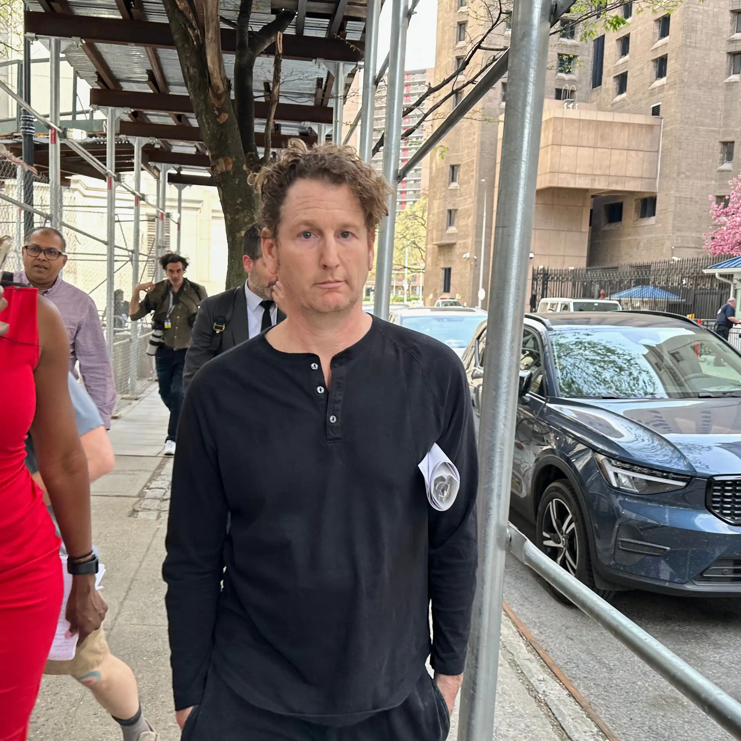 A man stares blankly ahead while walking and being videoed by a woman in a red dress.