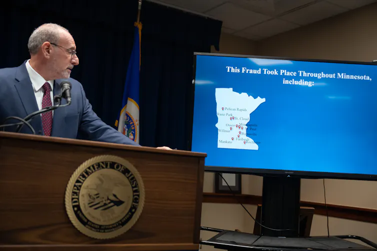 Un hombre de traje mira un mapa proyectado de Minnesota sobre un fondo azul en una pantalla junto a su podio.