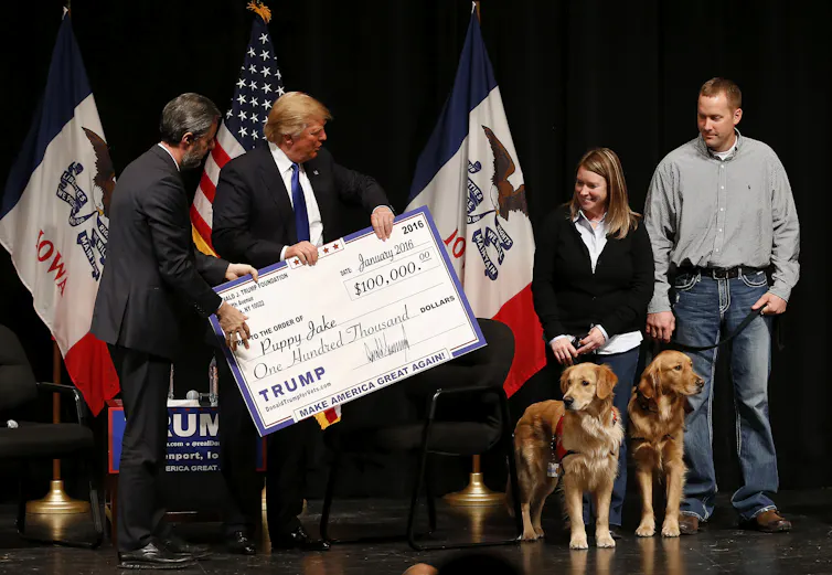 Donald Trump holds an oversized check and hands it to a woman holding the leashes for two dogs.