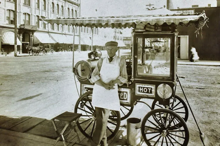Black and white photo: a man leans against a popcorn cart on the street.