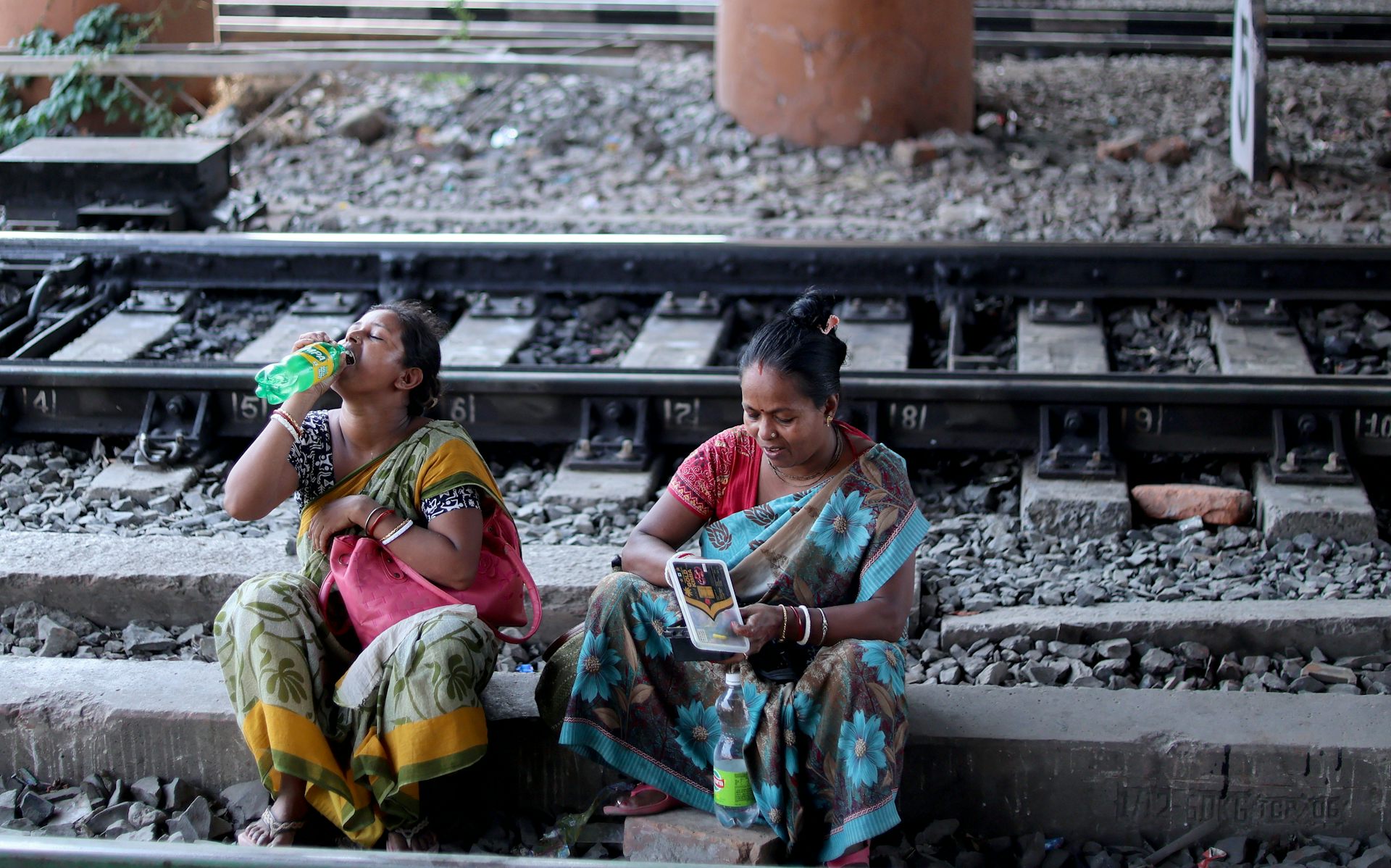 Two women sit on a train track, one of them drinking water.