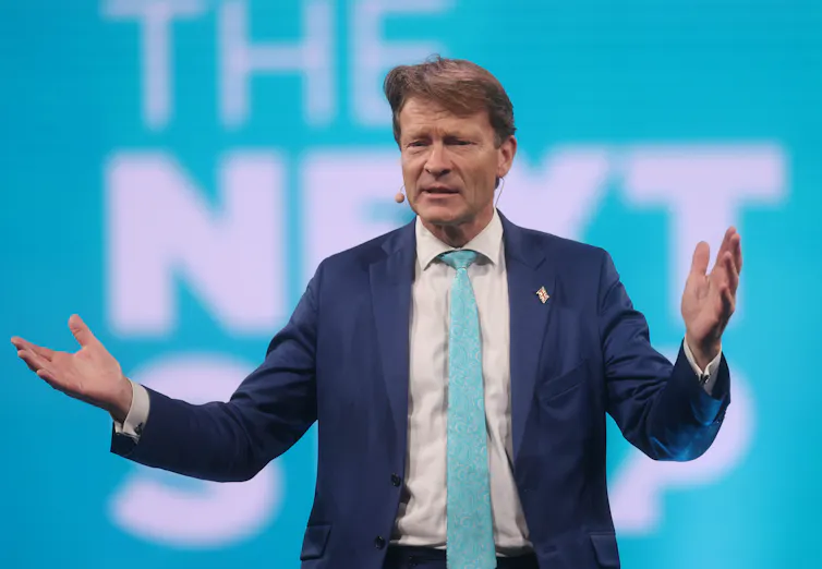 richard tice in suit and tie standing addressing an audience in front of a turquoise backdrop.