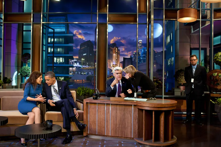 Un hombre, Obama, sentado en un sofá habla con una mujer mientras tras una mesa otro hombre, Jay Leno, habla con otra mujer.