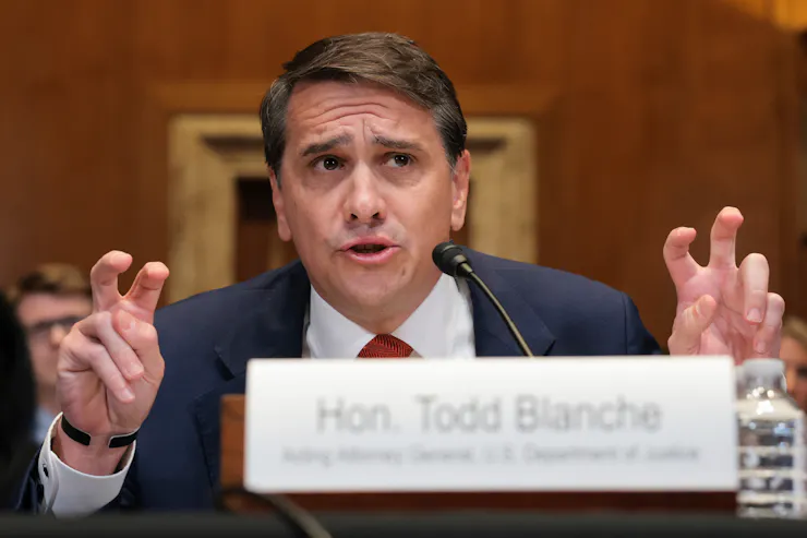 A man talking at a table behind a name plate, gesturing with his fingers.