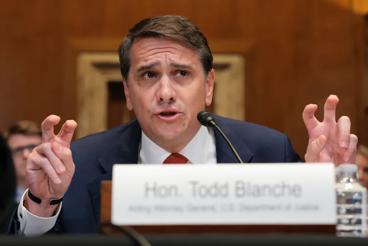 A man talking at a table behind a name plate, gesturing with his fingers.