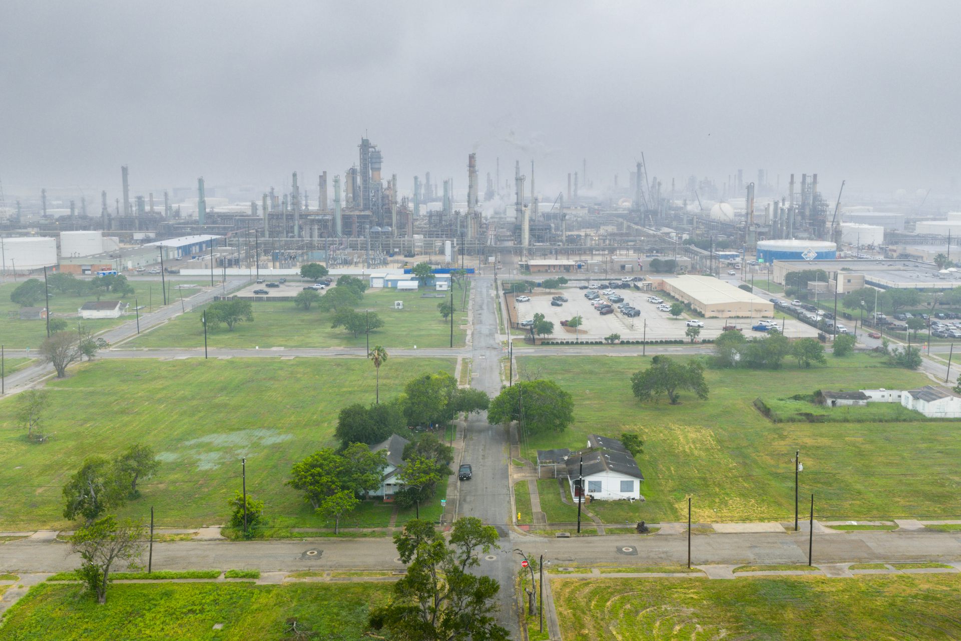 A neighborhood with grassy lawns and trees is in the foreground, with a massive industrial complex in the background.