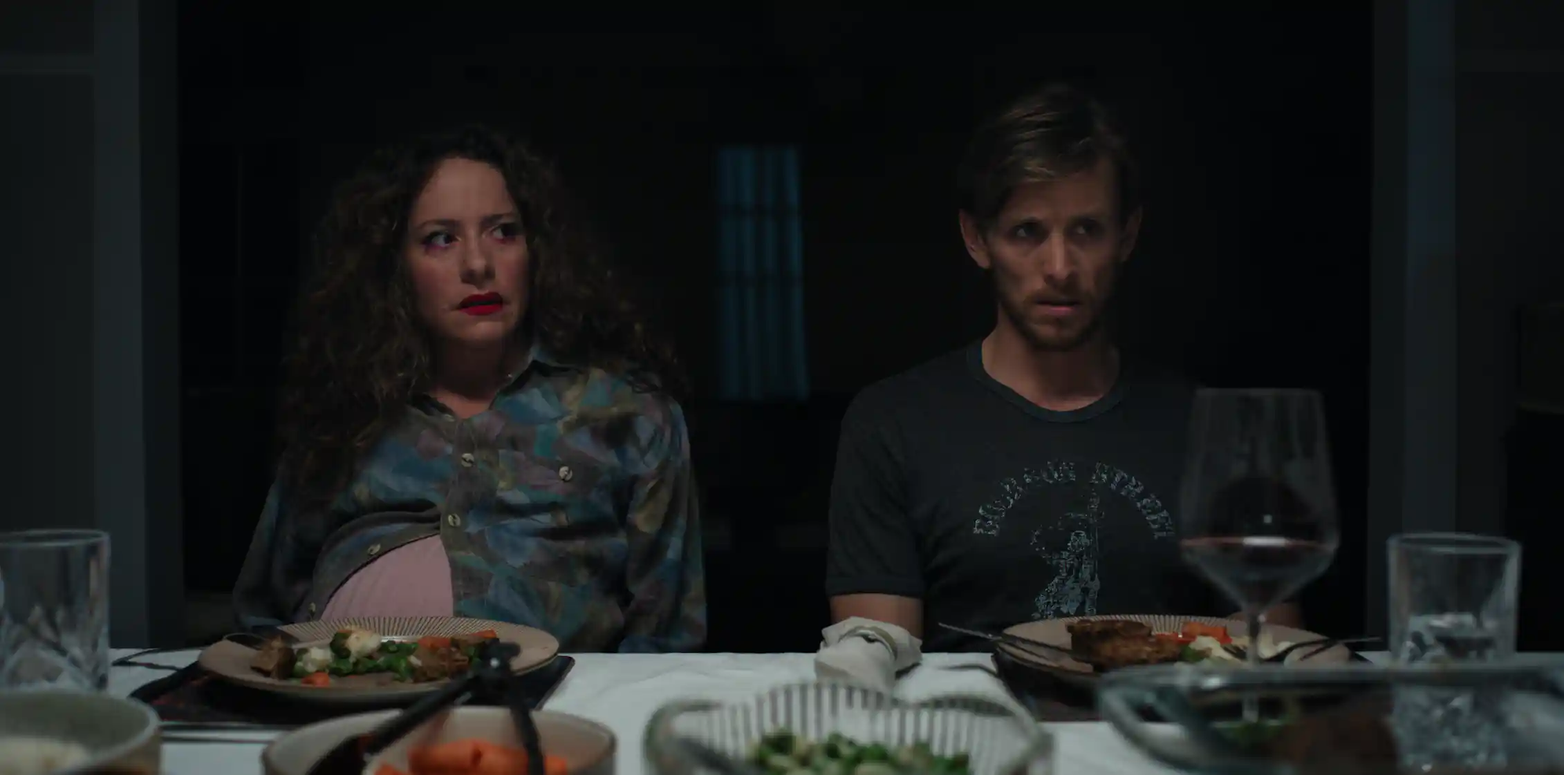 A woman and man sit at a dining table, in a dark room, with solemn expressions.