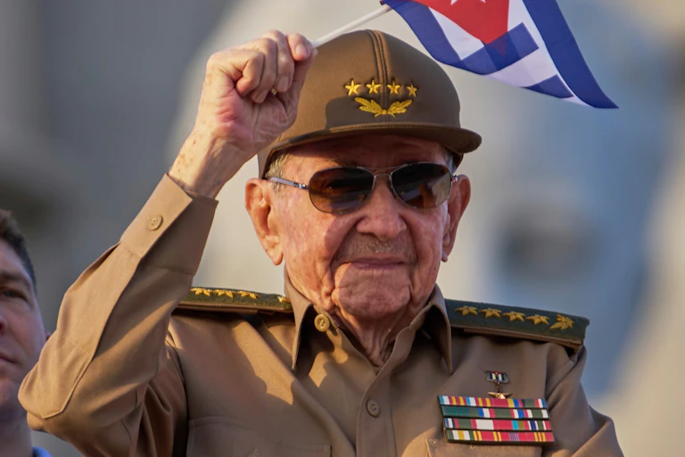 An old man in military uniform smiles and waves a small Cuban flag