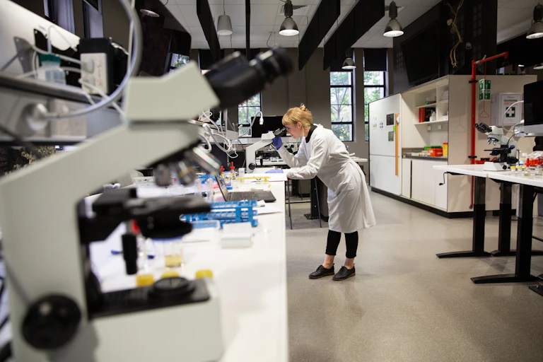 A young person wearing a lab coat looks in a microscope in a lab.