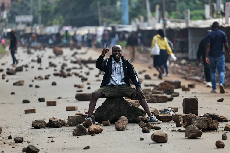a man sits on pile of bricks in road littered with rocks amid fuel price protest.