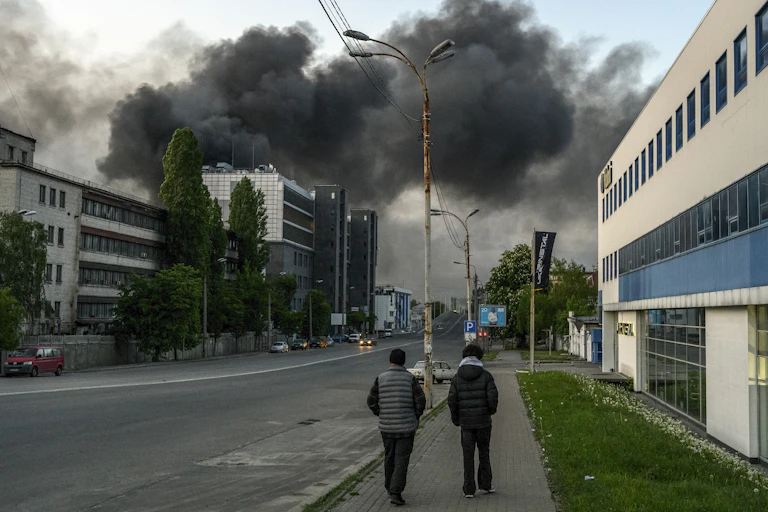 Local residents walk along a sidewalk as smoke rises over a street in Kyiv.