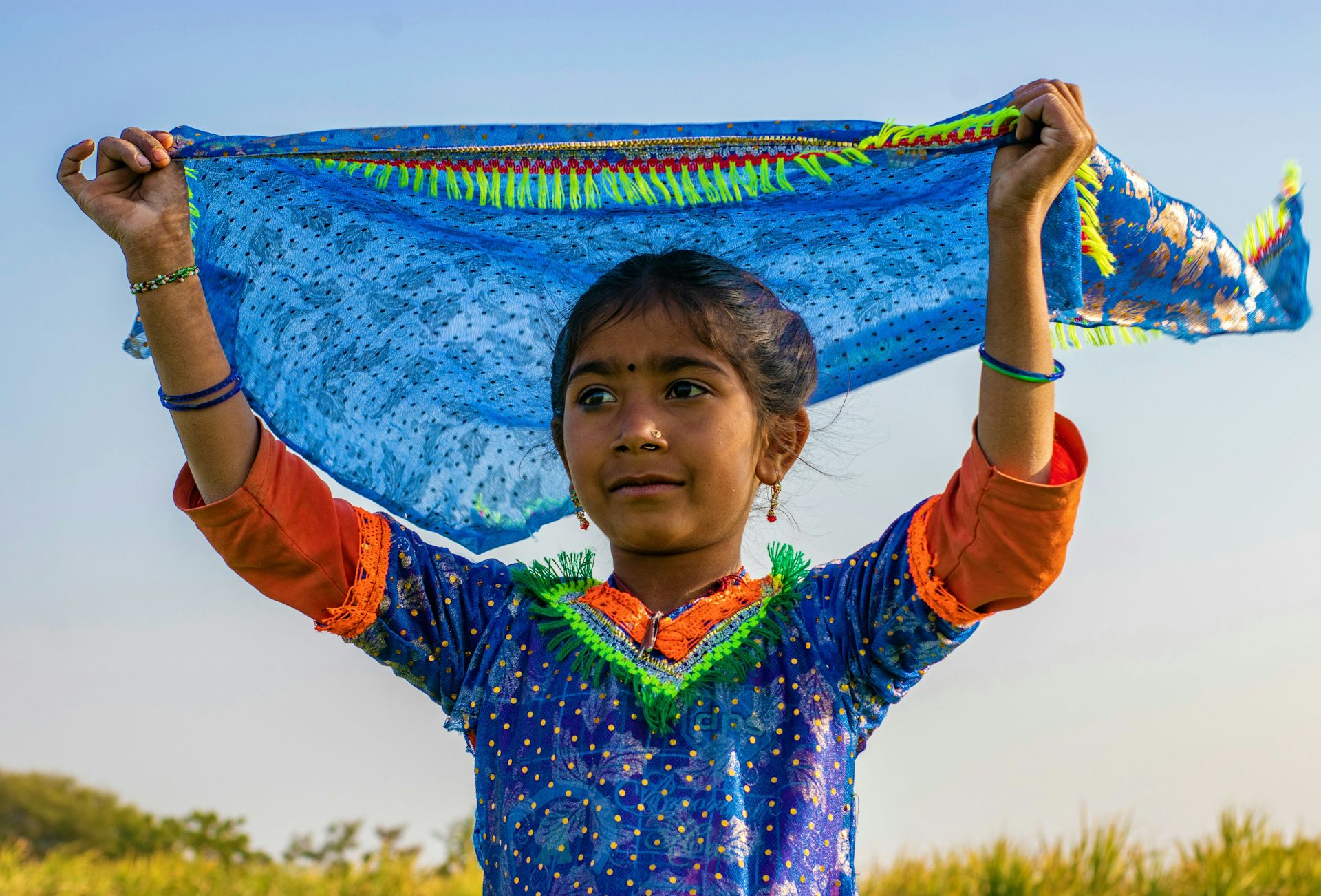 Una chica con un vestido colorido sostiene un pañuelo sobre su cabeza ondeando al viento.
