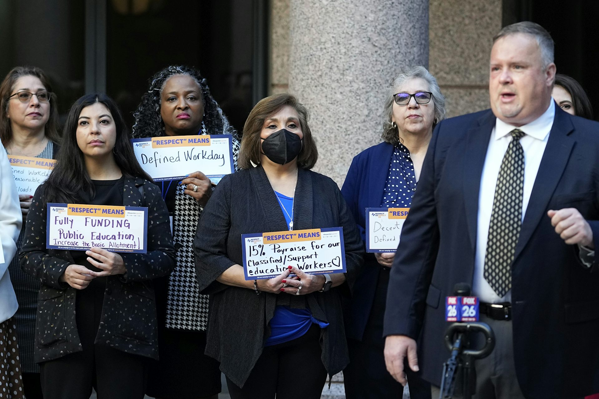People in face masks surround a speaker at a microphone while holding cardboard signs with slogans including 'defined workday' and 'fully funded public education.'