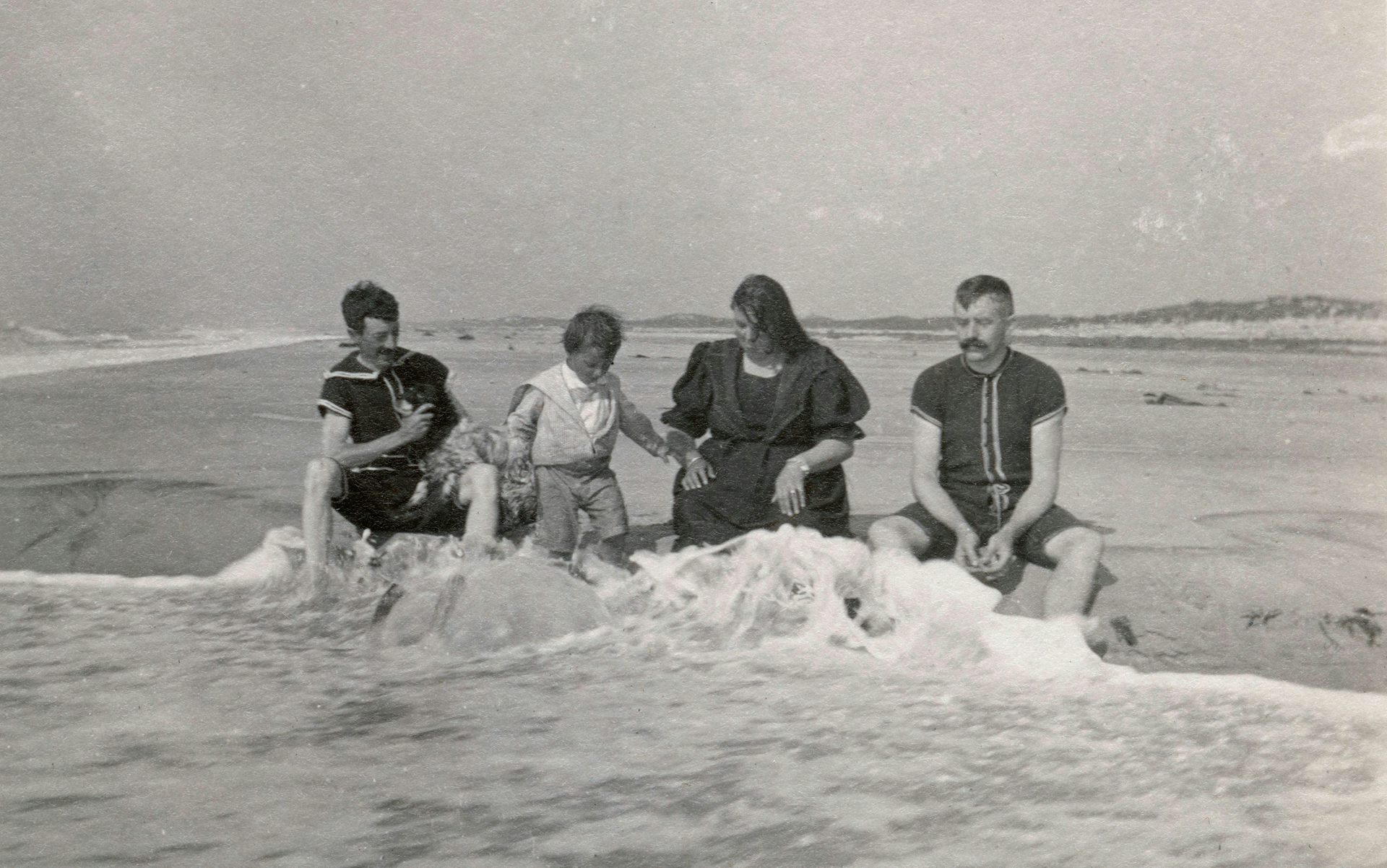 A family in old fashioned long bathing suits sit in the sea