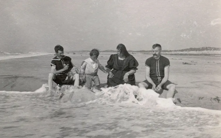 A family in old fashioned long bathing suits sit in the sea