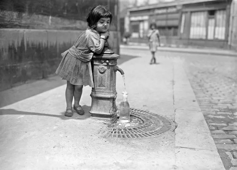 A young girl in Paris leaning on a public water fountain in 1921 while a bottle fills under the spout.