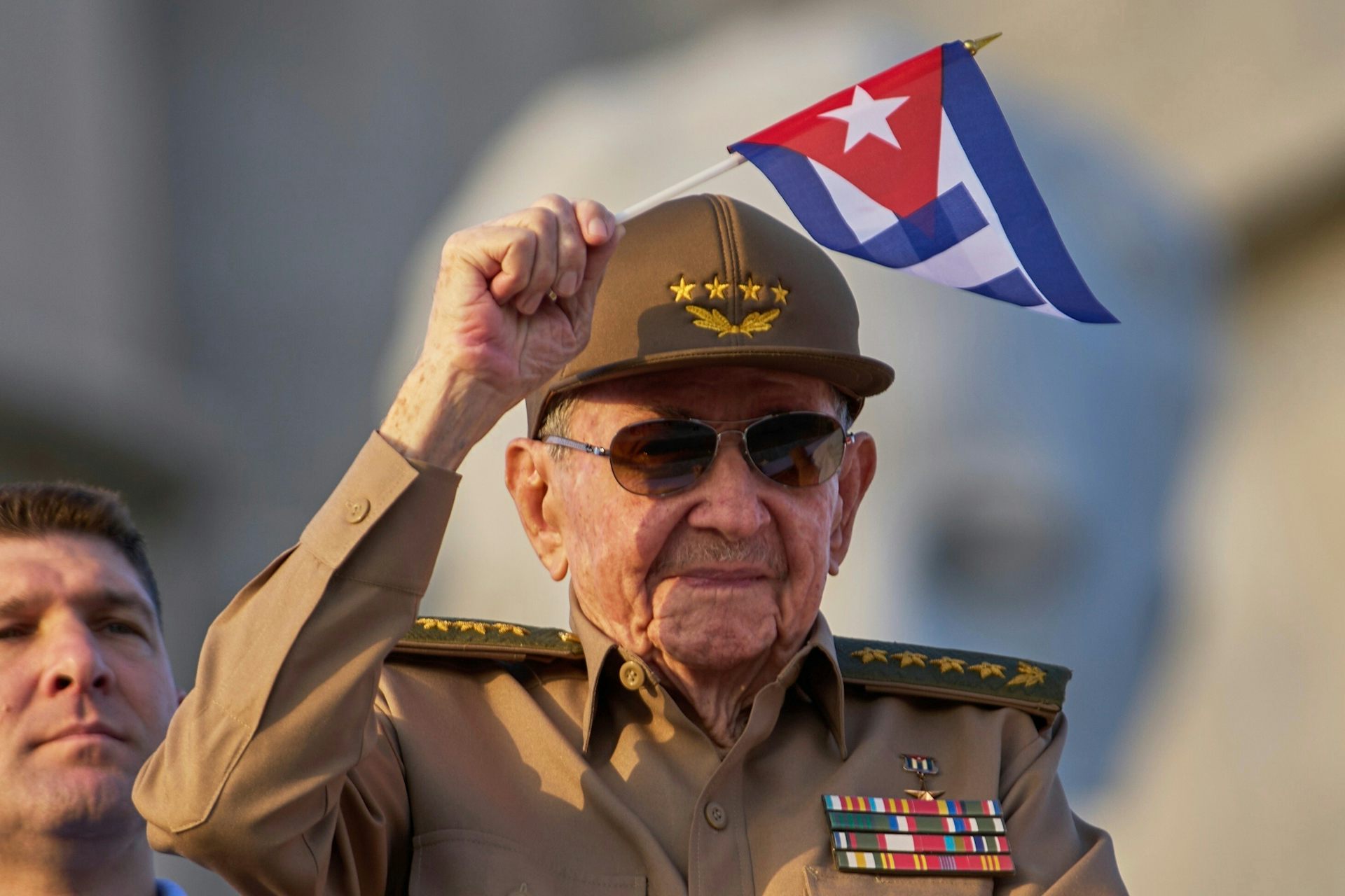 An old man in military uniform smiles and waves a small Cuban flag