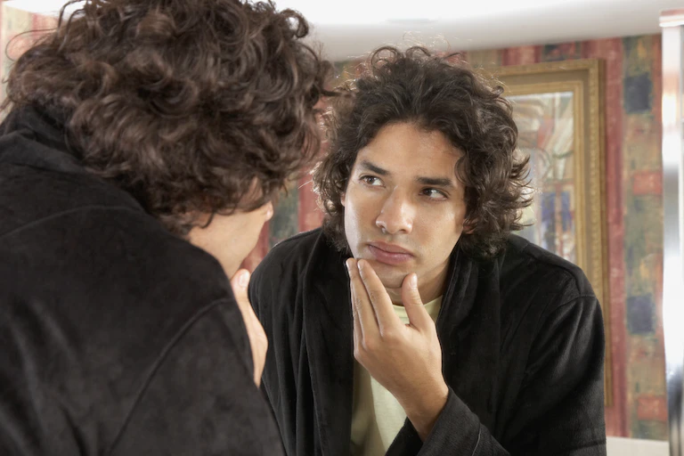 Young man with curly hair examining his face in the mirror.