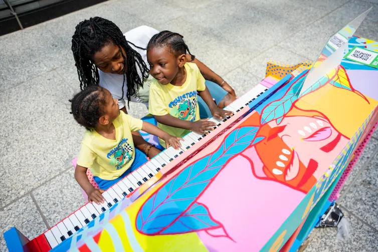 Two young Black girls and an accompanying adult sit playing a colourful outdoor piano.