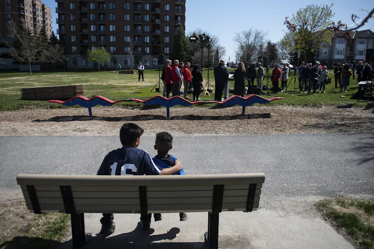 Se ve al niño mayor con su brazo alrededor del niño más pequeño, sentado en un banco del parque y mirando a los adultos en el parque y el área de juegos.