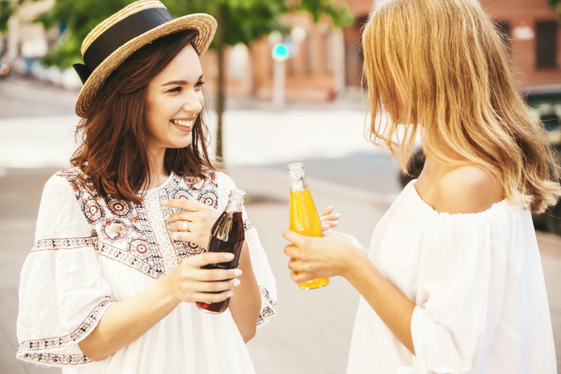 Two young women smile at each other, while one holds a bottle of coke or pepsi and the other a bottle of fanta or orange juice.