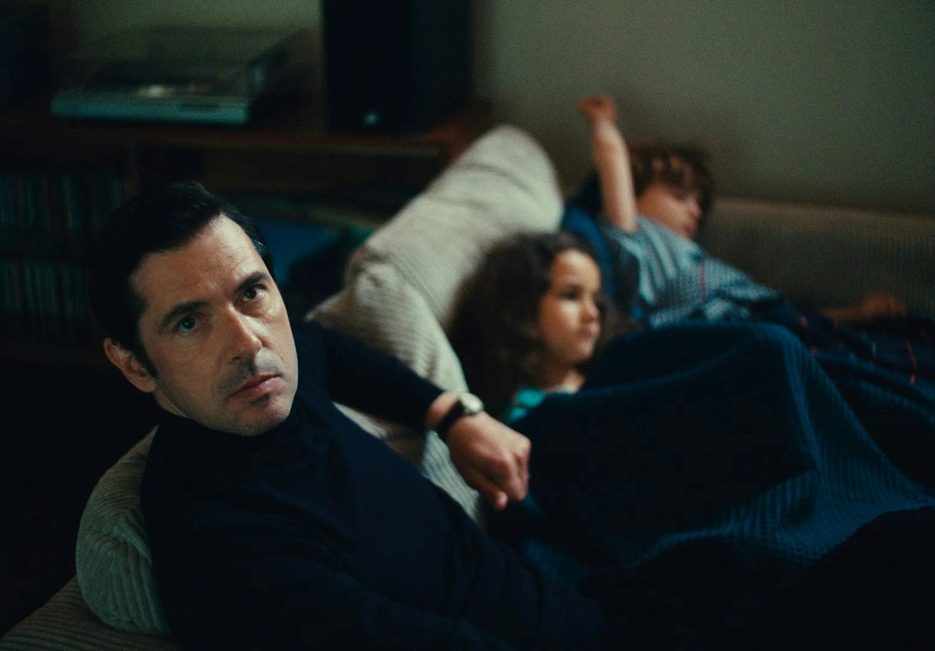 Image tirée du film, Melvil Poupaud sur un canapé regardant la caméra, les deux enfants du personnage qu'il joue sont à côté de lui.