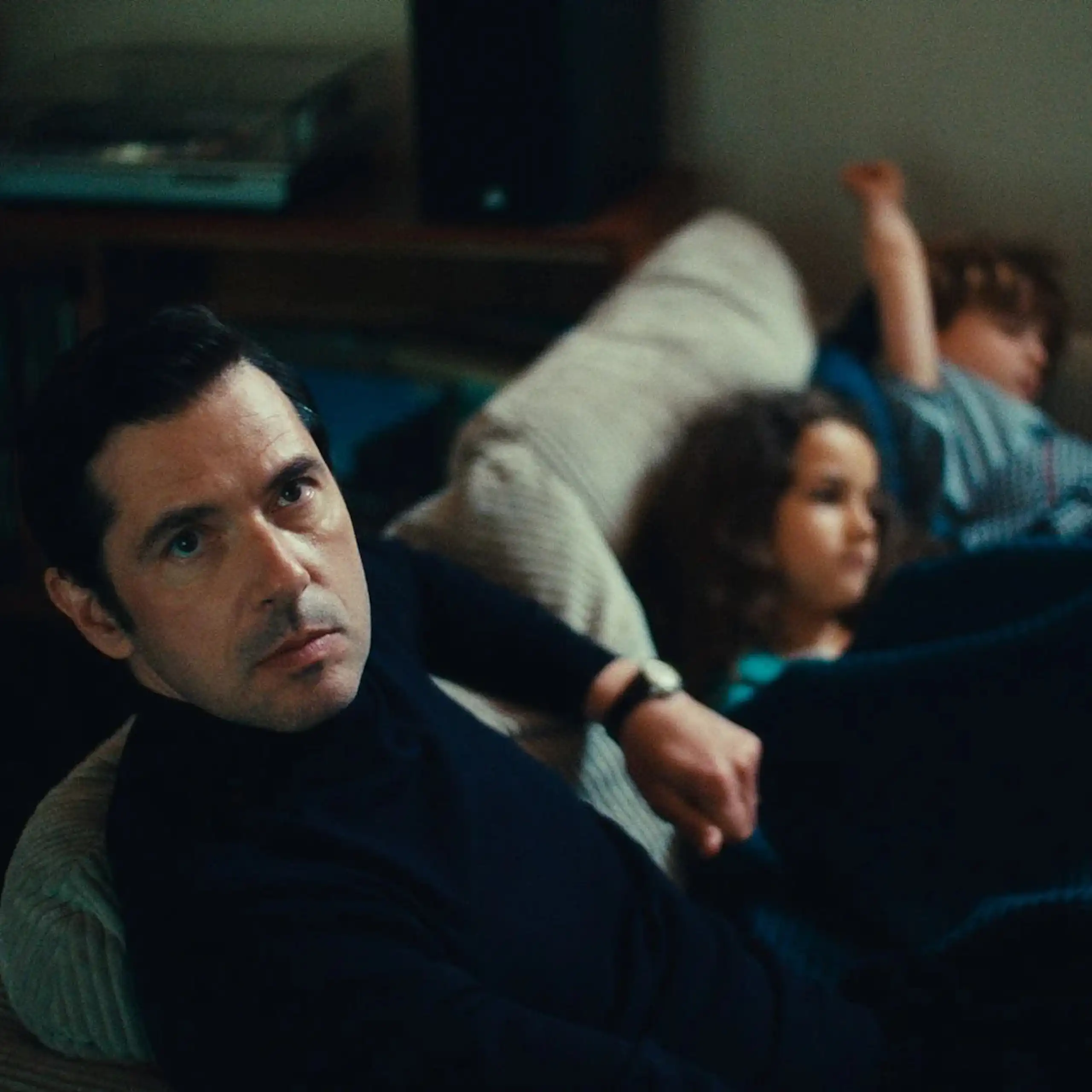 Image tirée du film, Melvil Poupaud sur un canapé regardant la caméra, les deux enfants du personnage qu'il joue sont à côté de lui.