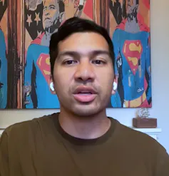 A young dark-haired man in a khaki jumper speaking to the camera.