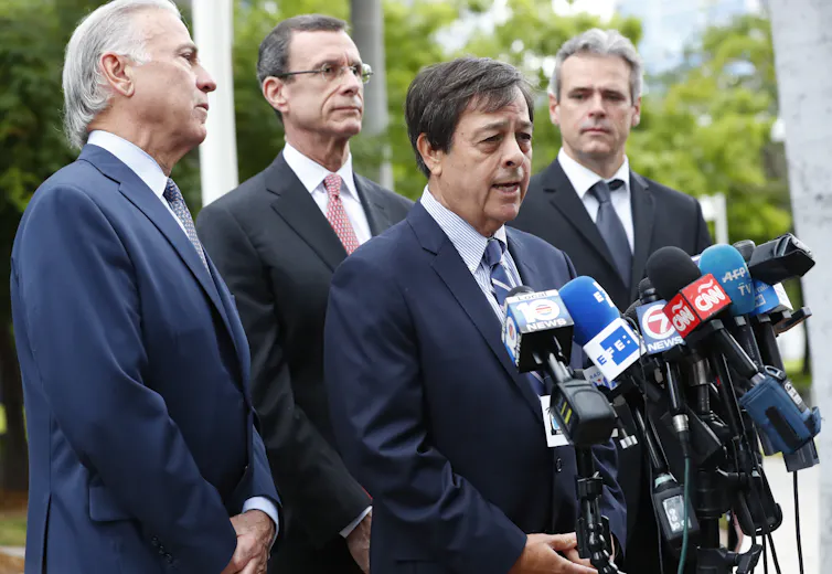 Four men in suits stand by a lot of microphones, with one of them in front, speaking.